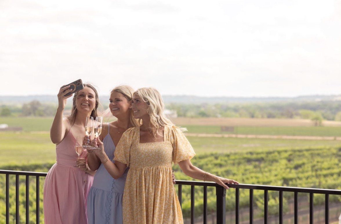 Three friends taking a selfie with wine on a balcony above the vineyards